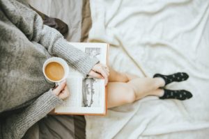 photo of a woman's torso and legs reading a book and drinking coffee