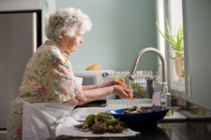 elderly woman with short white curly hair washing vegetables at the sink near the window