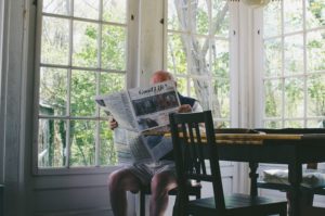 elderly man sitting at a kitchen table reading a newspaper in a sunny room full of windows