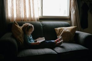 picture of a little girl reading a book on the couch near a window